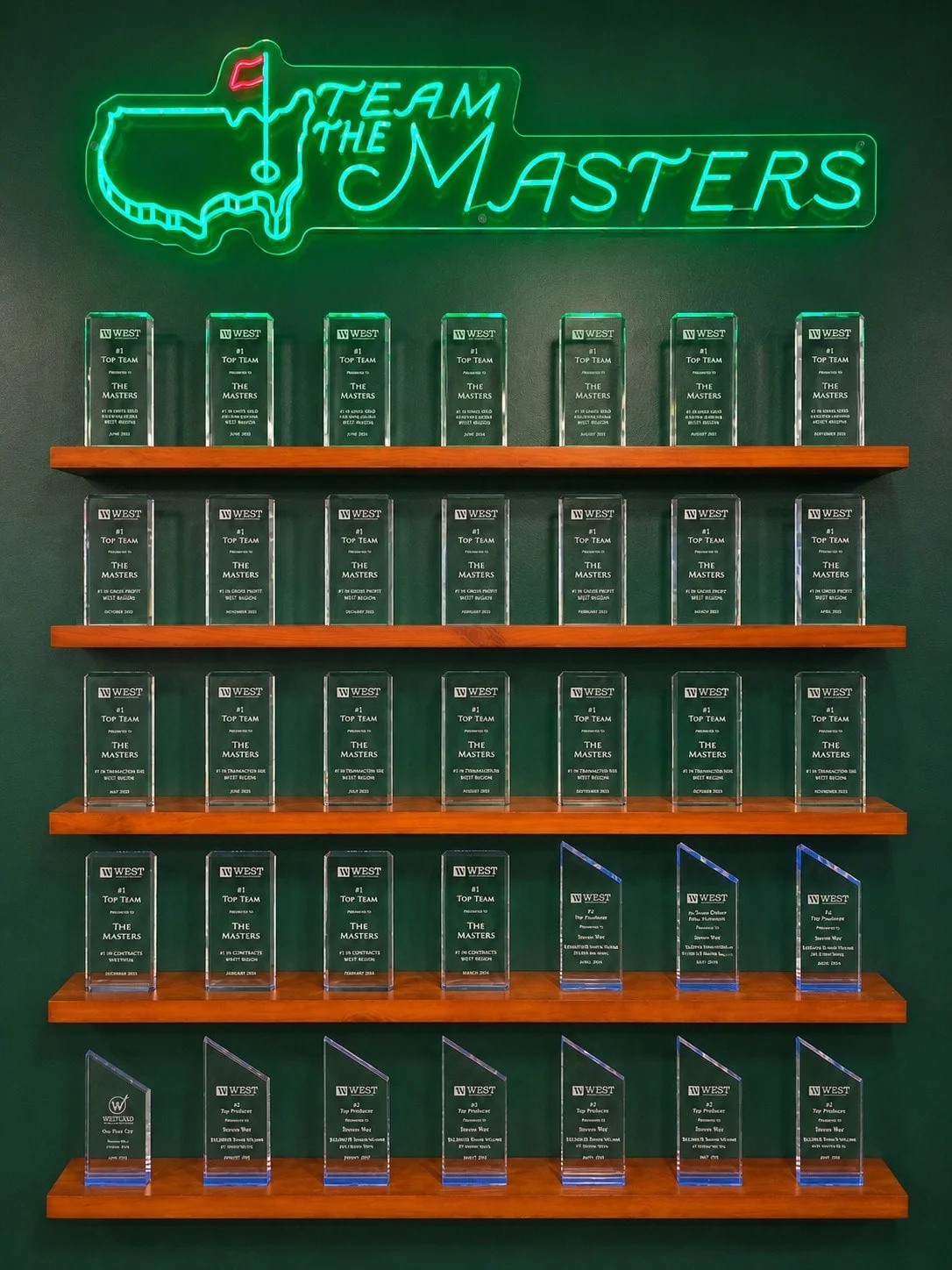 Glass award plaques on five shelves beneath The Masters neon sign in the office